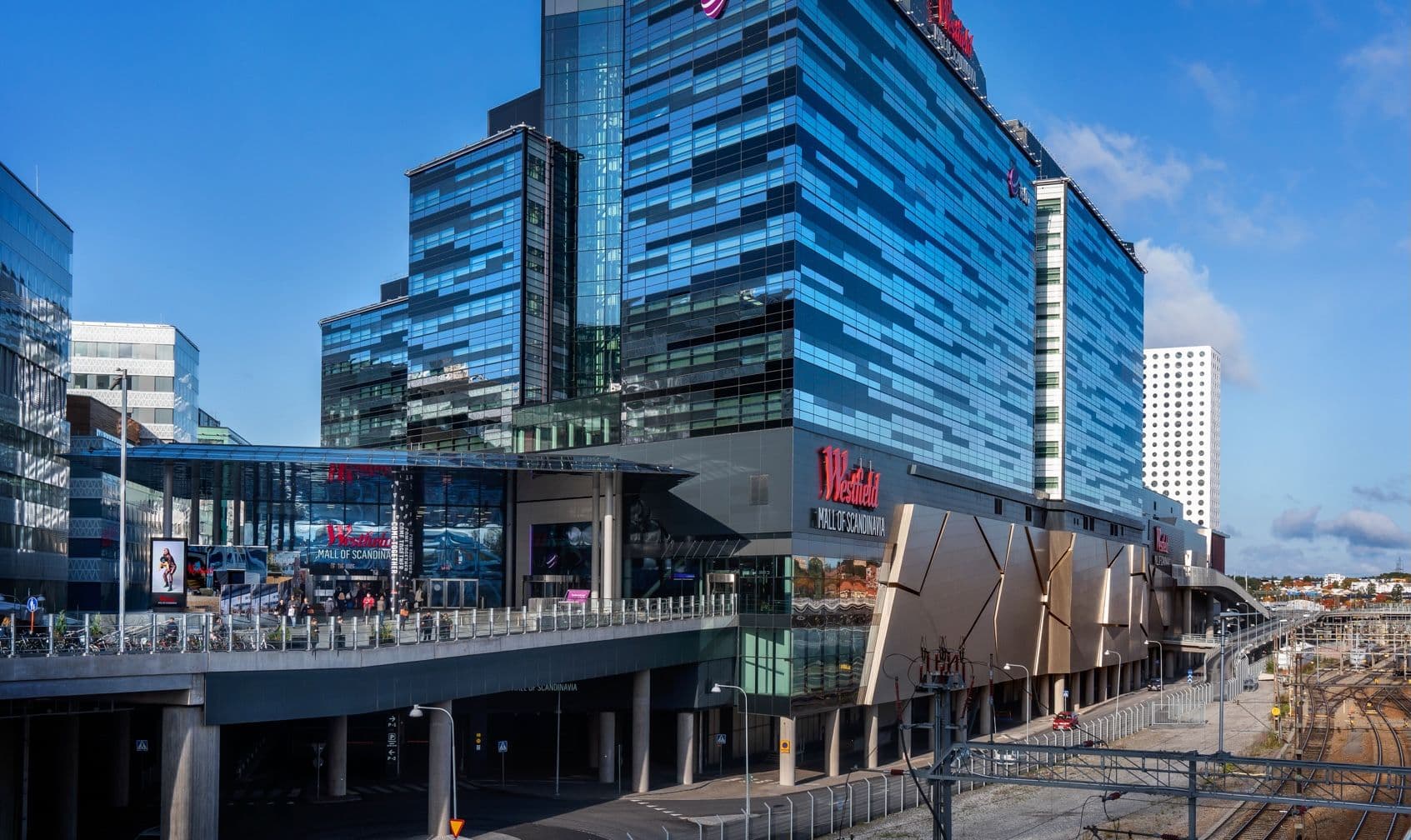 Huvudentrén till Mall of Scandinavia. Till höger om entrén är ett stort kontorshus med mycket glas. På fasaden sitter en röd skylt där det står "Westfield".